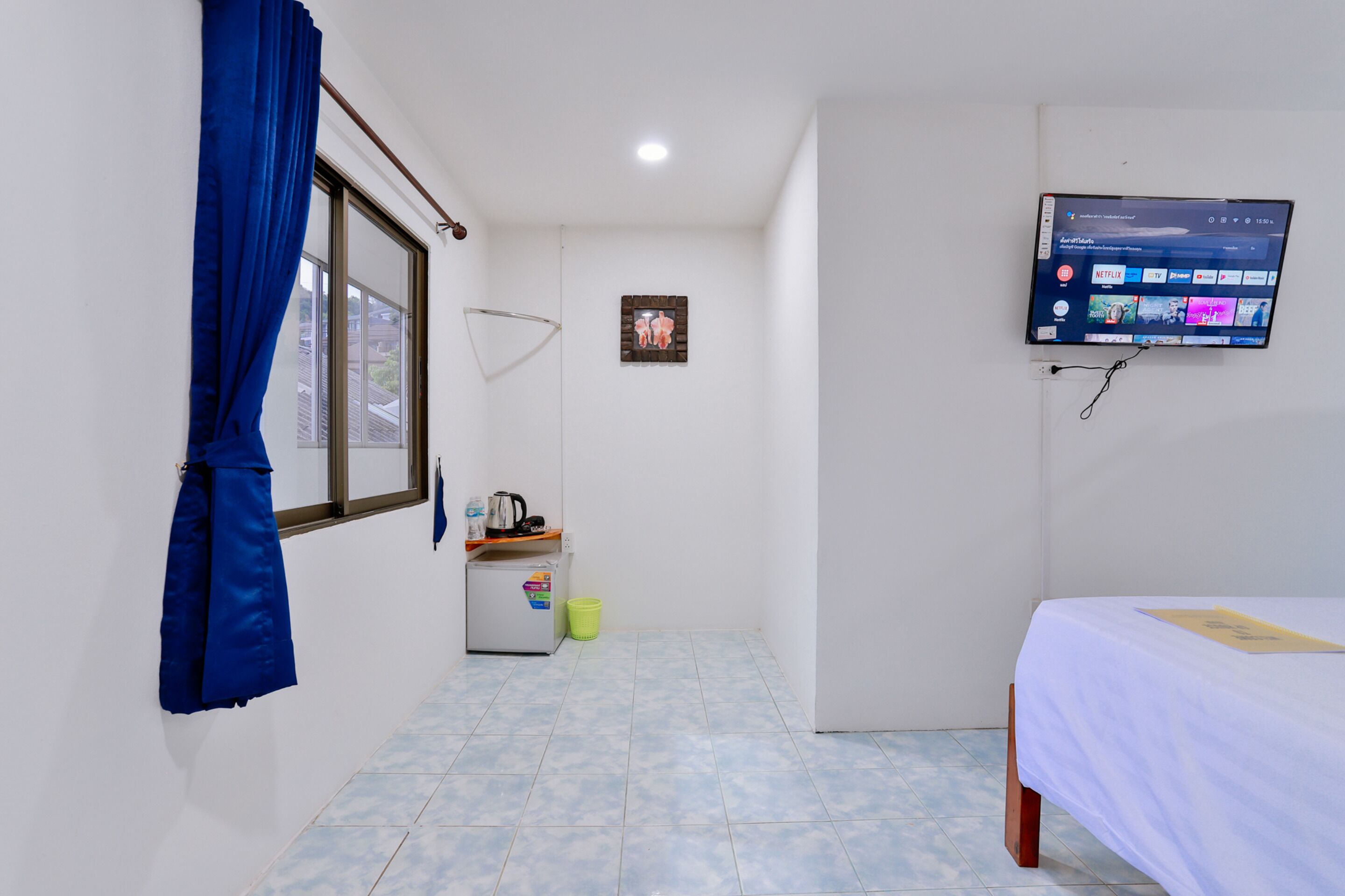 A small, clean room with a bed, a TV mounted on the wall, a window with blue curtains, a small refrigerator, and a trash bin.