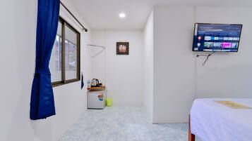 A small, clean room with a bed, a TV mounted on the wall, a window with blue curtains, a small refrigerator, and a trash bin.