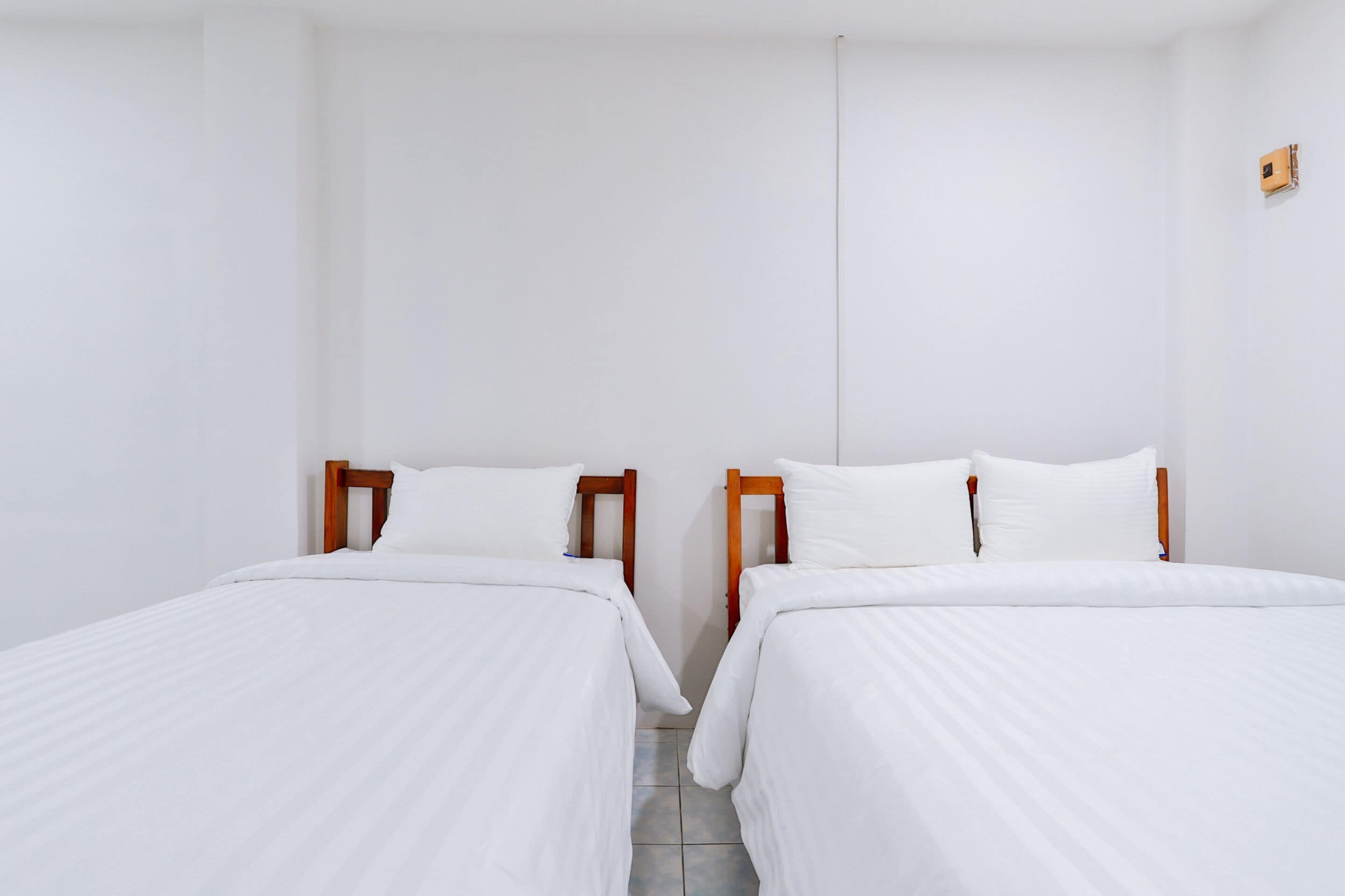 A clean, minimalist room with two single beds, wooden headboards, white bedding, and a small wall-mounted device.