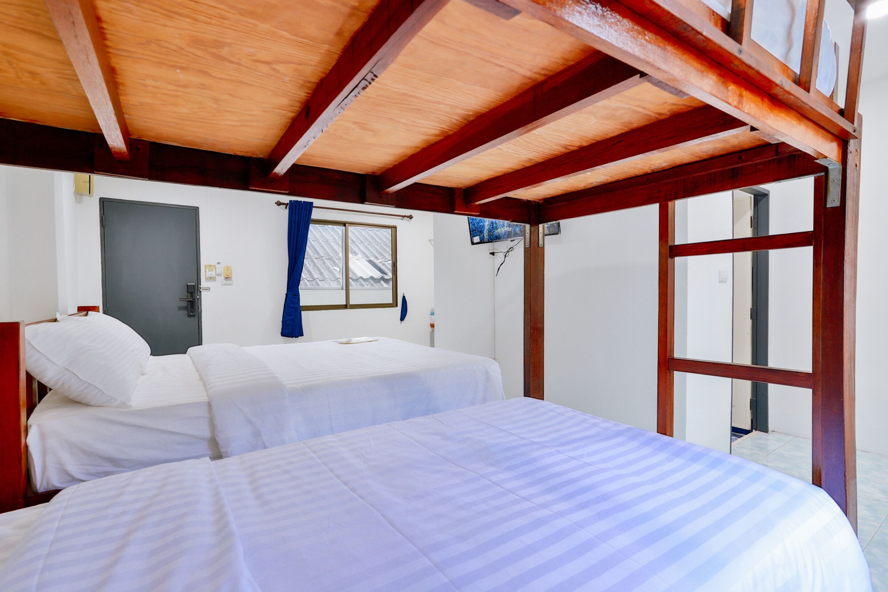 Habitación de hotel con dos camas, vigas de madera en el techo, una ventana con cortinas azules y una puerta.