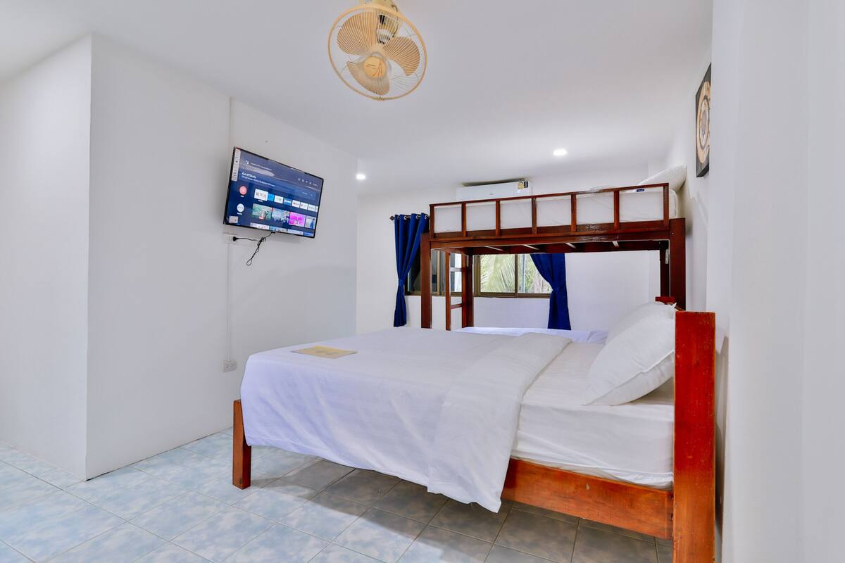 A bedroom with a wooden bed, white bedding, a ceiling fan, a mounted TV, and a window with curtains.