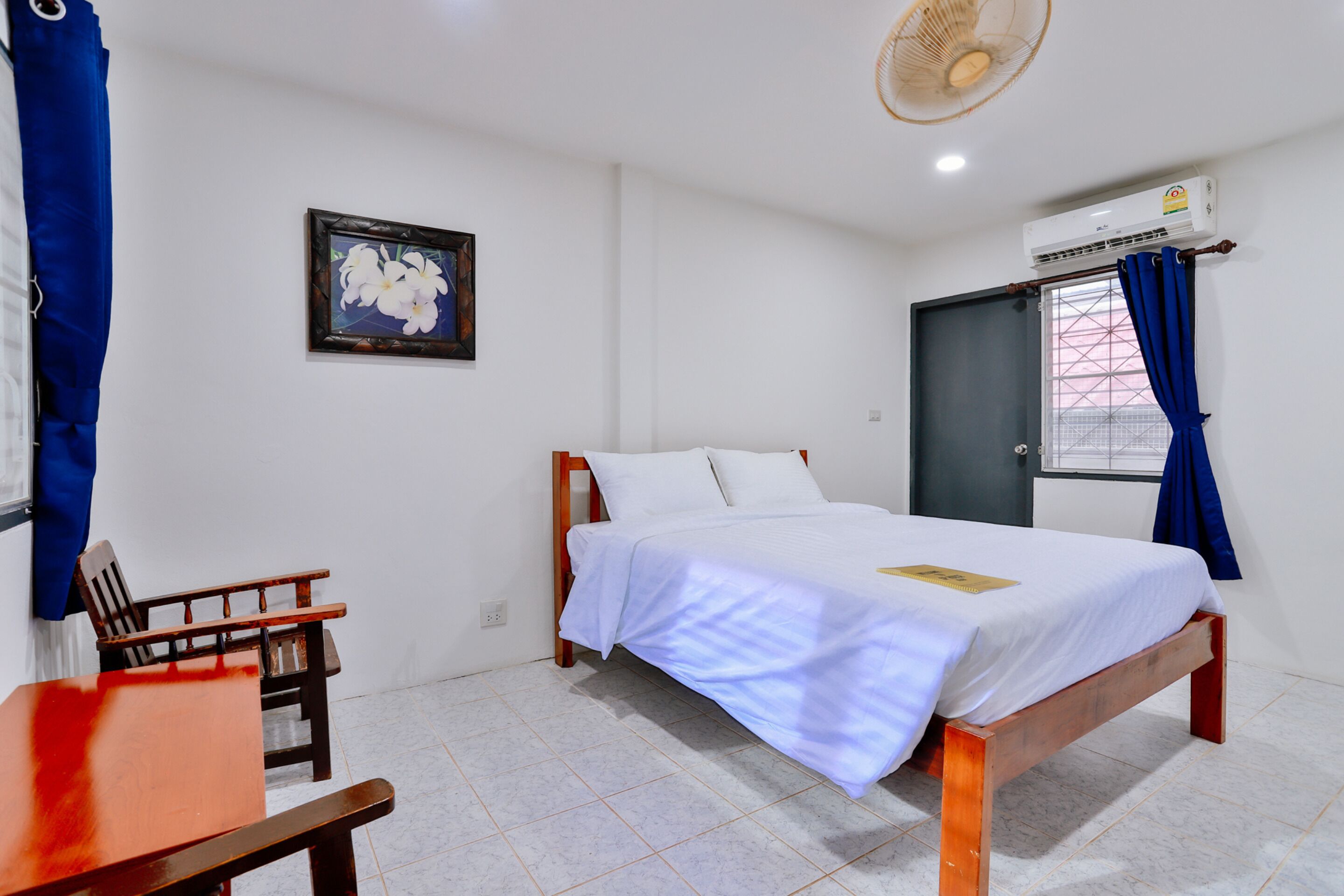 A bedroom with a bed, a chair, a table, a picture on the wall, and an air conditioning unit.