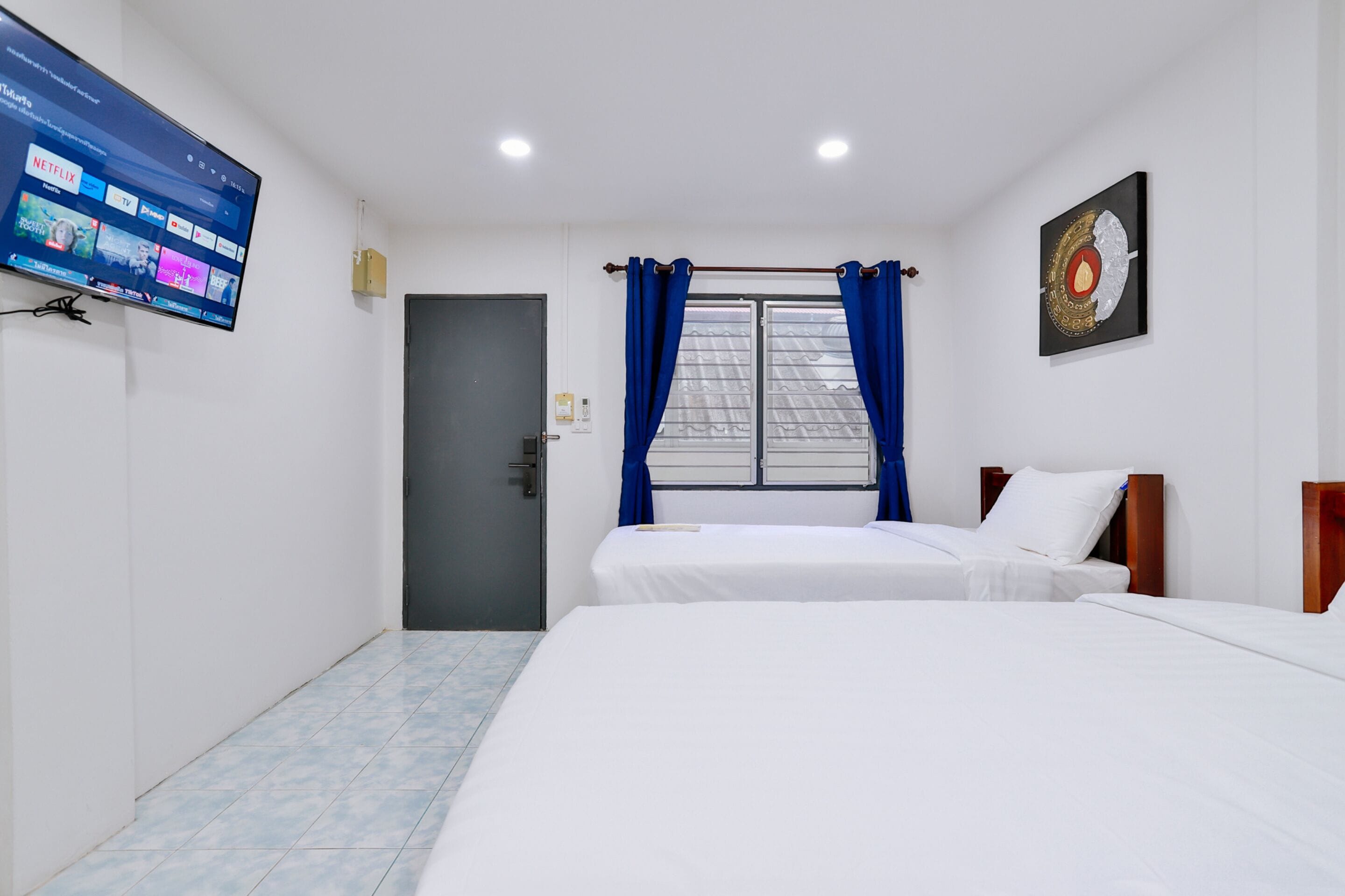 Habitación de hotel con dos camas, un televisor montado en la pared, una ventana con cortinas azules y una puerta.
