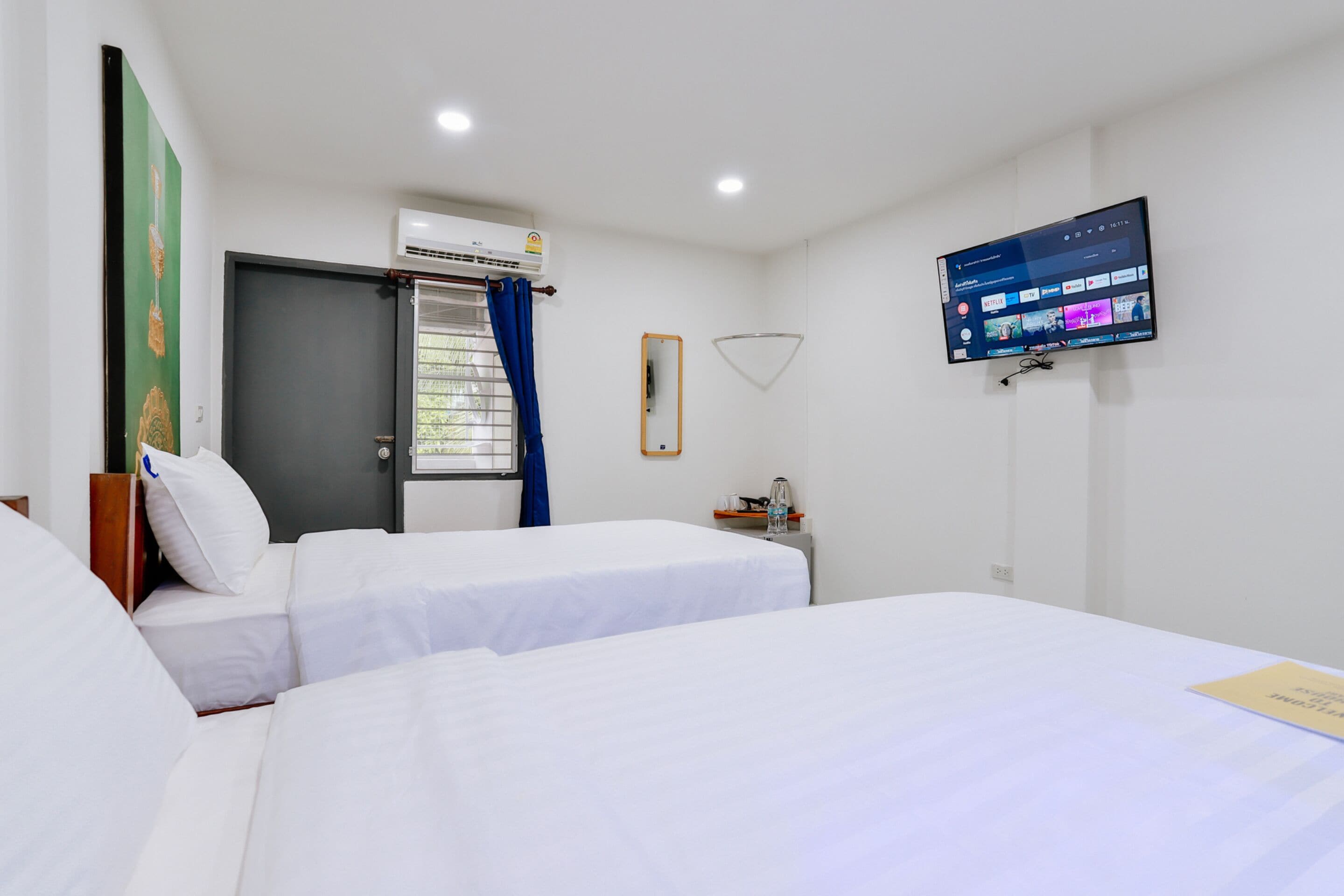 A hotel room with two beds, a TV mounted on the wall, an air conditioning unit, and a window with curtains.