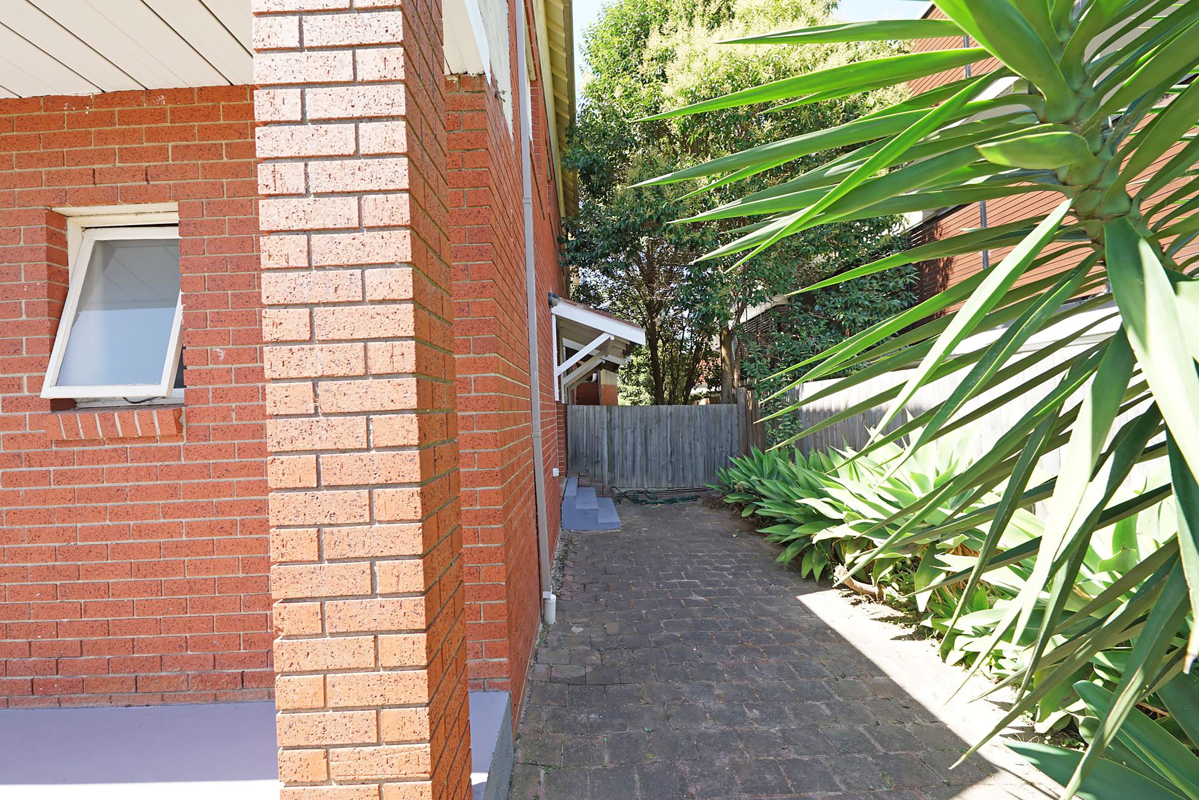 A narrow brick pathway leading to a wooden gate, surrounded by lush green plants and trees.