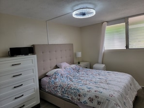 A bedroom with a bed, a dresser, a nightstand, a lamp, a ceiling fan, and a window with blinds.