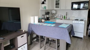 Una cocina compacta con microondas, estufa y gabinetes blancos. Hay una mesa con mantel azul y un televisor sobre un atril de madera.
