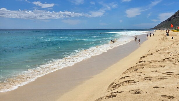 Una playa con cielos despejados, aguas tranquilas y huellas en la arena.