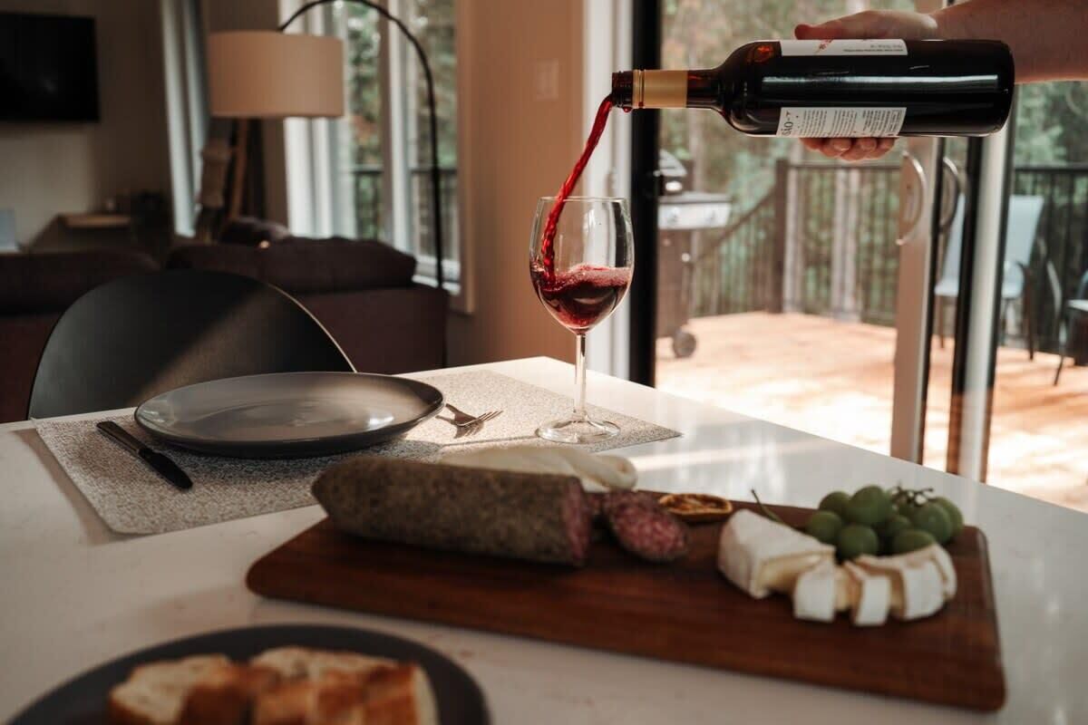 Une table avec une bouteille de vin rouge dont on verse le contenu dans un verre, une planche à fromages avec différents types de fromages et un couteau, et une assiette avec du pain.
