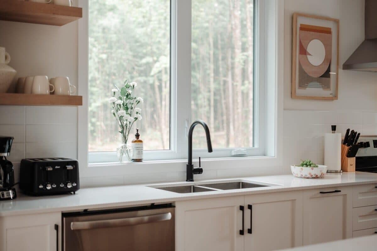 A modern kitchen with white cabinets, a black faucet, a toaster, and a window with a view of trees.