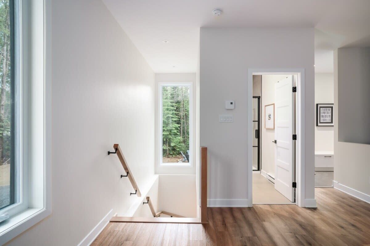Un interior moderno con una escalera de madera, vista a árboles por una ventana y un pasillo que conduce a otra habitación.
