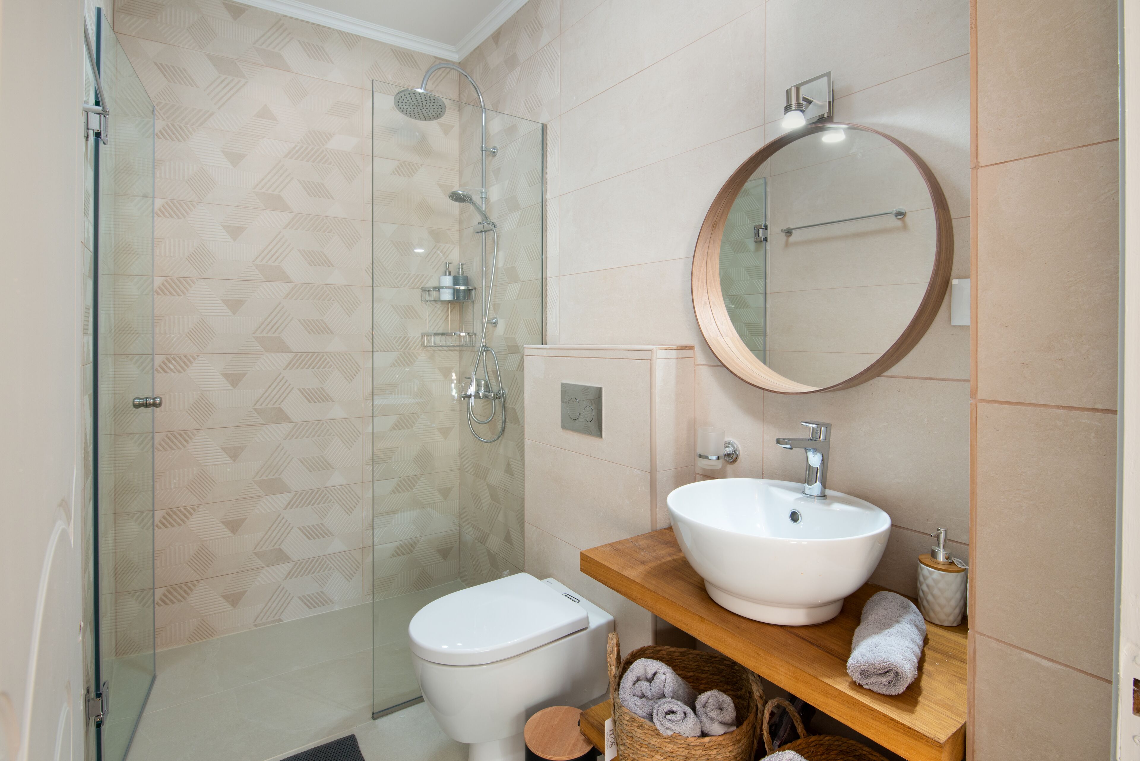 Un baño moderno con ducha de vidrio, inodoro blanco, tocador de madera con espejo redondo y lavamanos blanco.