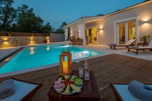 A poolside area with a wooden deck, lounge chairs, a table with a fruit platter, and a lantern.