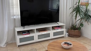 A living room with a TV on a white stand, a round wooden table with a remote, and a potted plant. - Centrally Located Coastal Getaway – Walk to Beaches, Cafes & More (Miami)