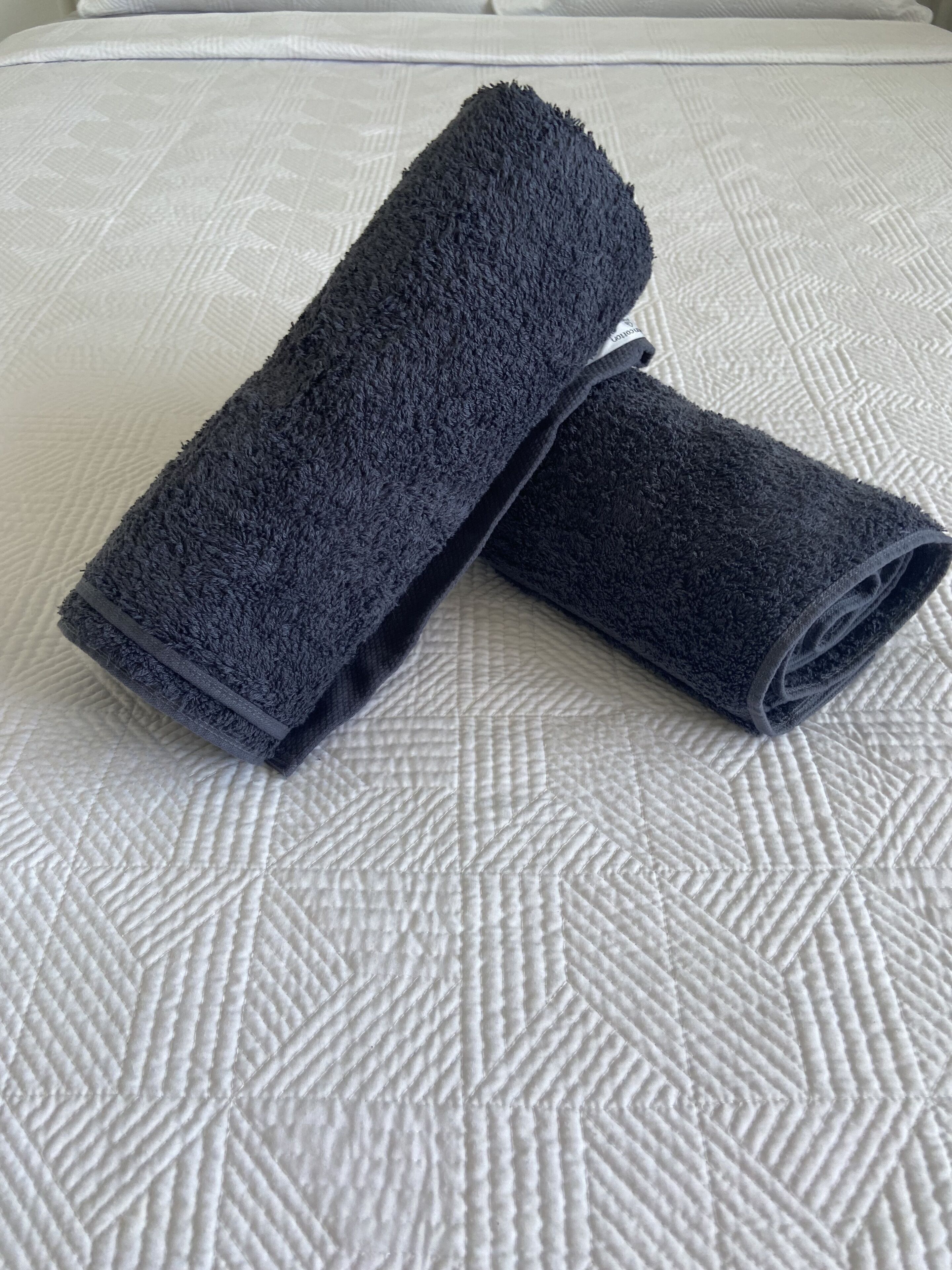 Ein Bett mit einer gesteppten weißen Decke und zwei gerollten, dunkelblauen Handtüchern.