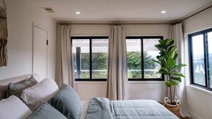Een slaapkamer met een bed, twee ramen met gordijnen, een kamerplant en uitzicht op groen.