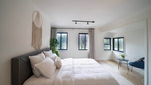 Een moderne slaapkamer met een groot bed, twee ramen, een klein tafeltje met een plant en een hangend macramé kunstwerk.