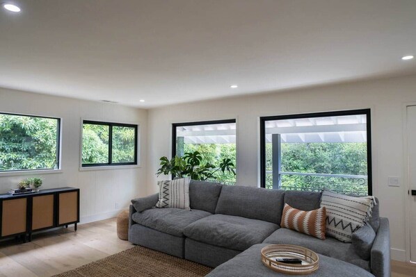 Uma sala de estar moderna com um sofá seccional cinza, uma mesinha lateral e uma janela grande com vista para o verde.