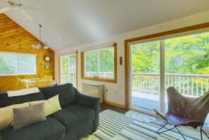 Un salon chaleureux avec un plafond en bois, un canapé vert, une table à manger avec des chaises et une vue sur une terrasse en bois, visible à travers de grandes fenêtres.
