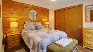 Um quarto com cabeceira de madeira, cama com colcha xadrez, duas luminárias de cabeceira, mesa de cabeceira, cômoda de madeira e um espelho na parede.