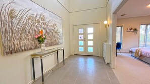 A hallway with a large abstract painting, a console table with a vase of flowers, and a bedroom with a bed and a window. - Spacious 7BRM 5-bathroom Rental House (Point Cook)