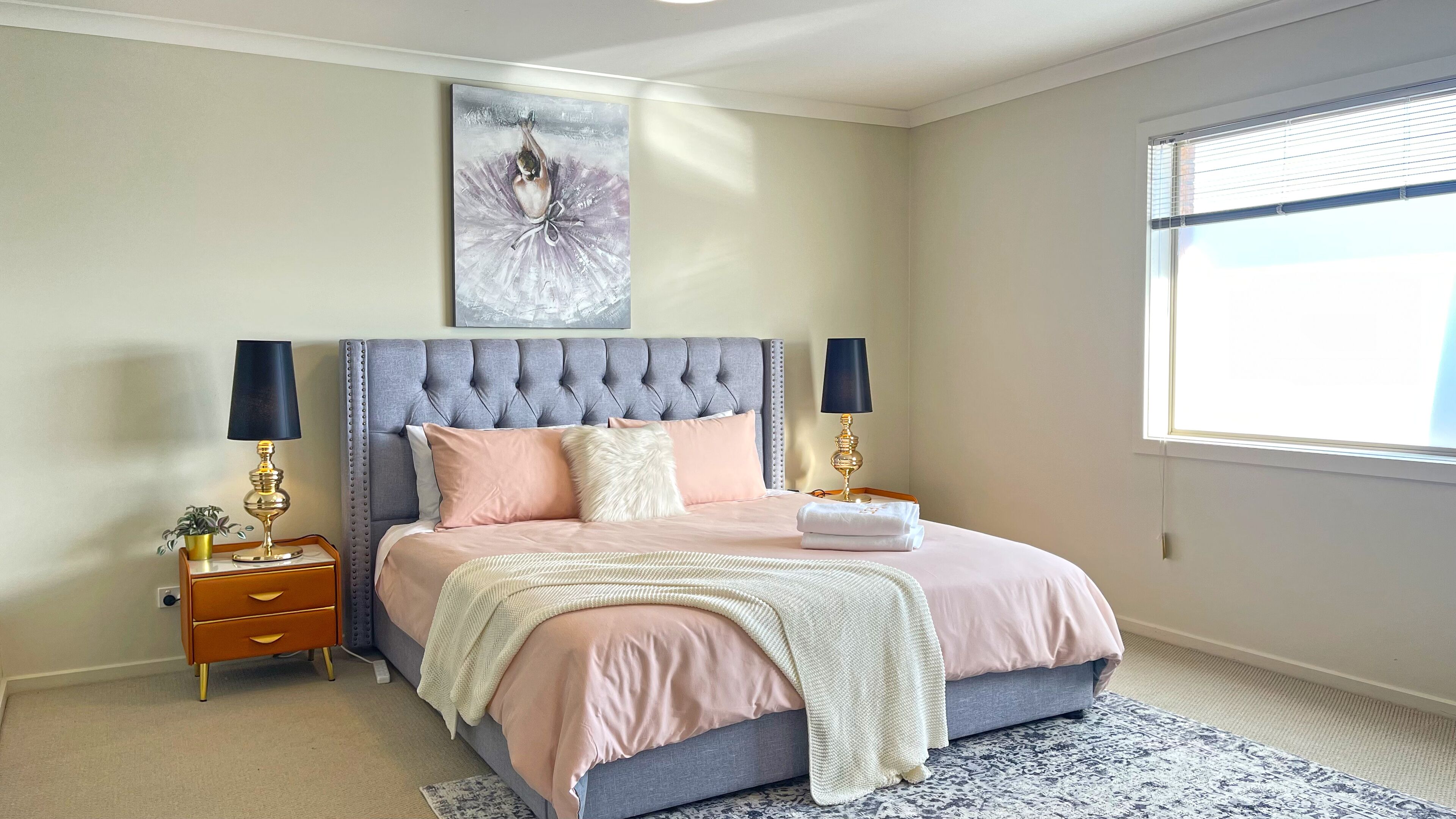 A bedroom with a tufted headboard, two bedside tables with lamps, a large window with blinds, and a painting above the bed.