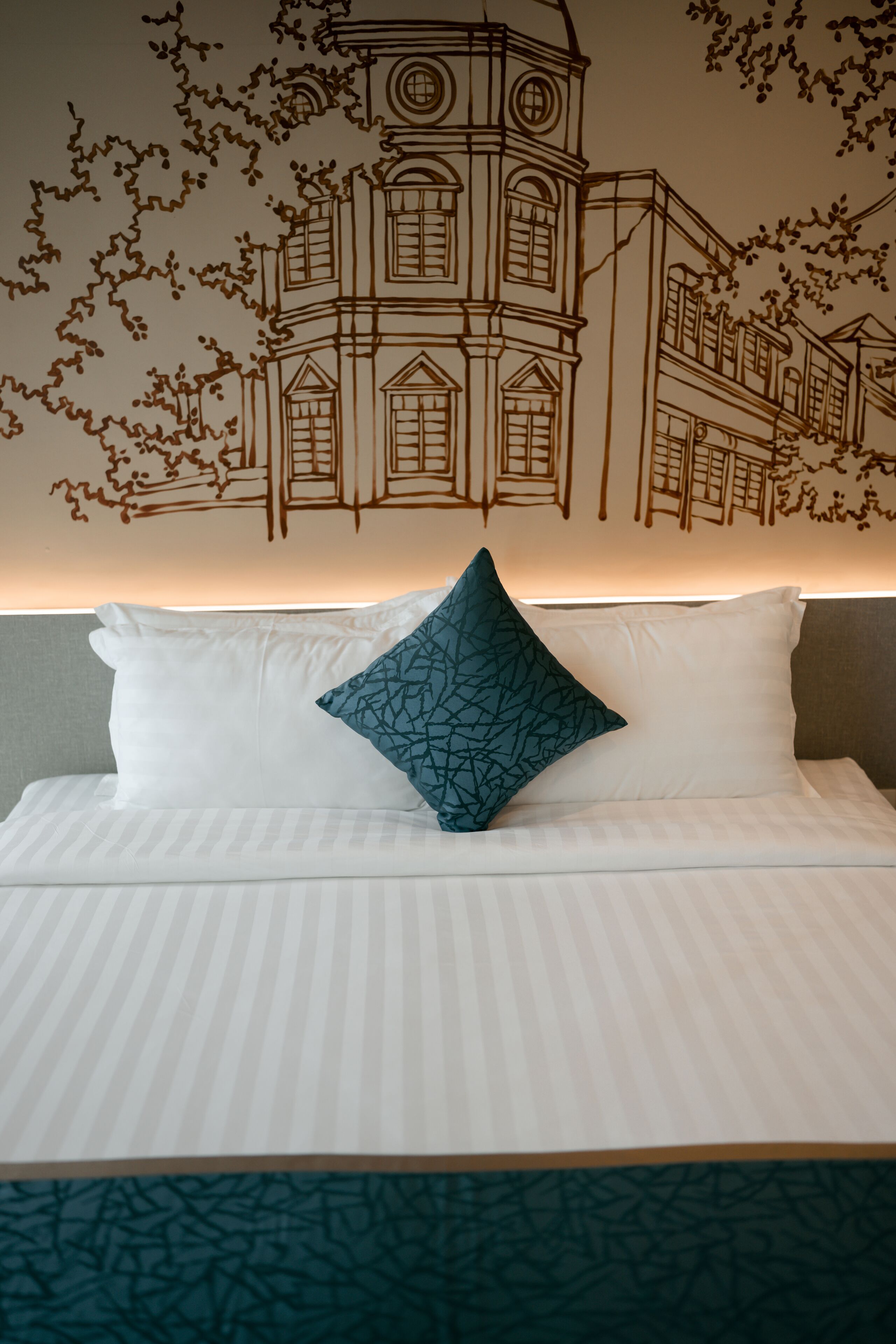 A neatly made bed with white linens and a single teal patterned pillow, set against a wall with a detailed mural of a building.