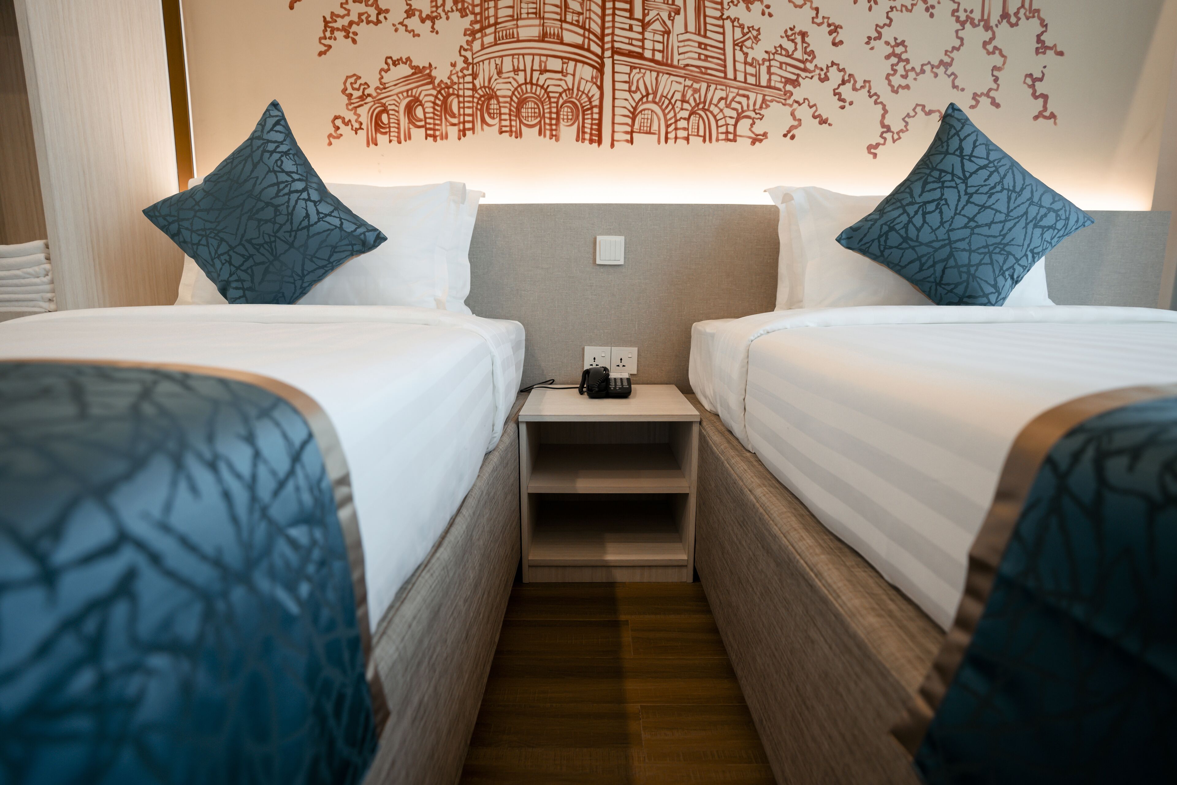 A hotel room with two beds, each with a patterned pillow and a headboard. There is a nightstand with a phone and a power outlet between the beds.