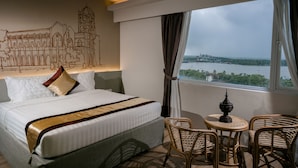 A hotel room with a large bed, a small table with chairs, and a view of a body of water through a window.
