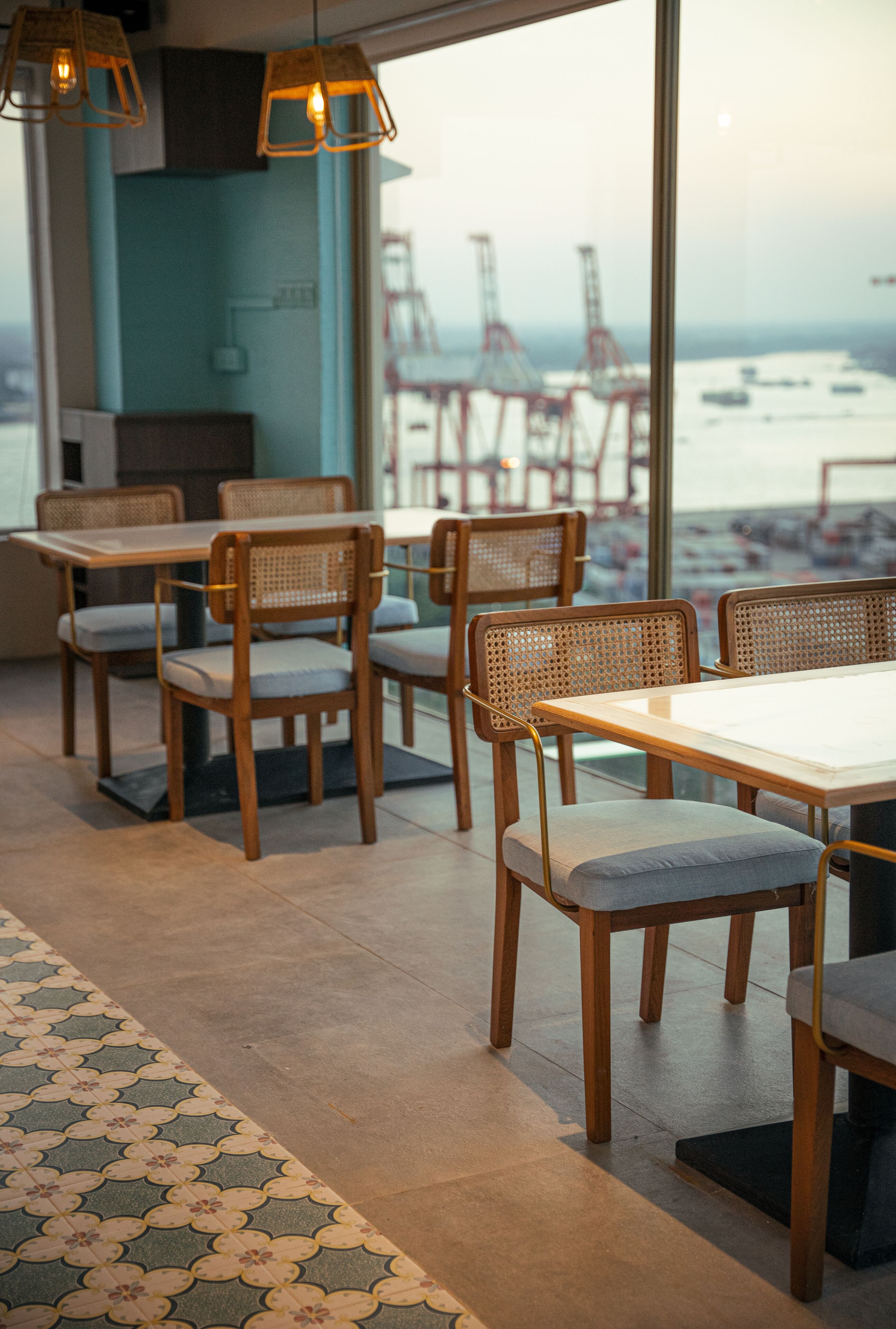 Un restaurant avec des tables et des chaises en bois, de grandes fenêtres donnant sur des grues et l’eau, et un sol à motifs.