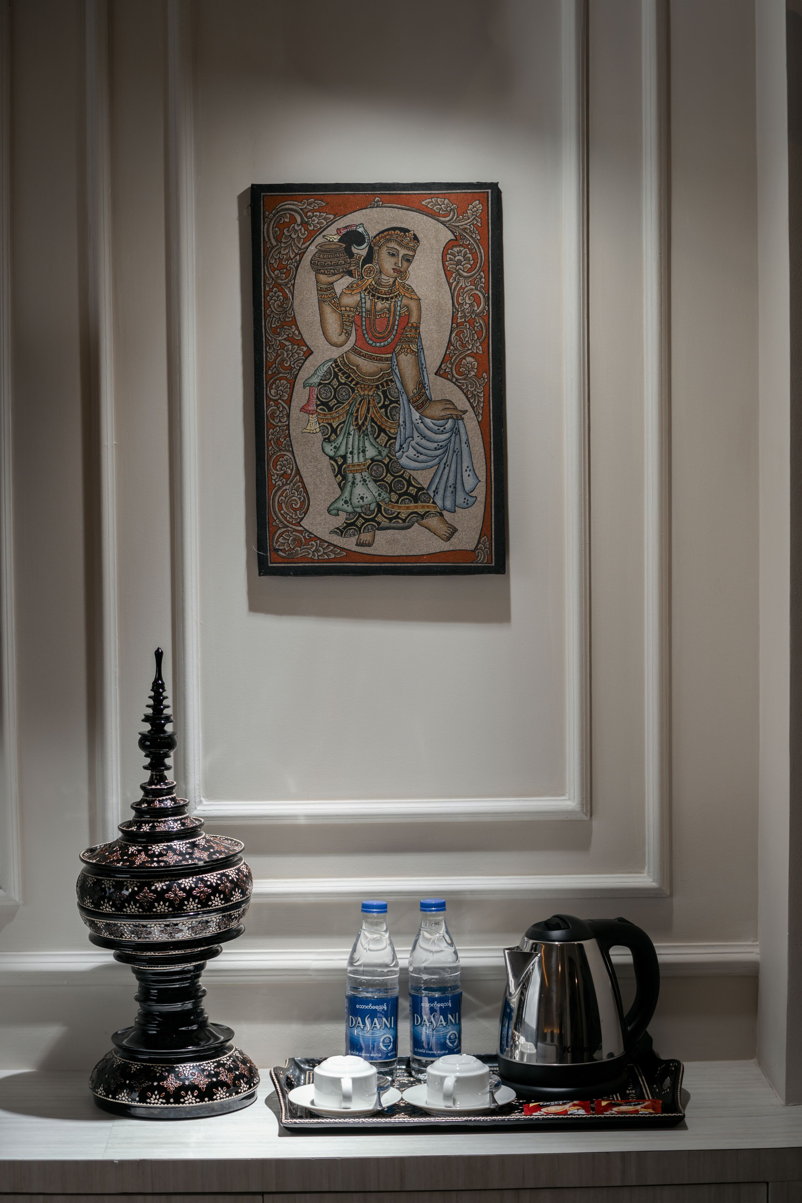A framed artwork on a wall, a decorative black object, and a tray with a water bottle, cup, and kettle.