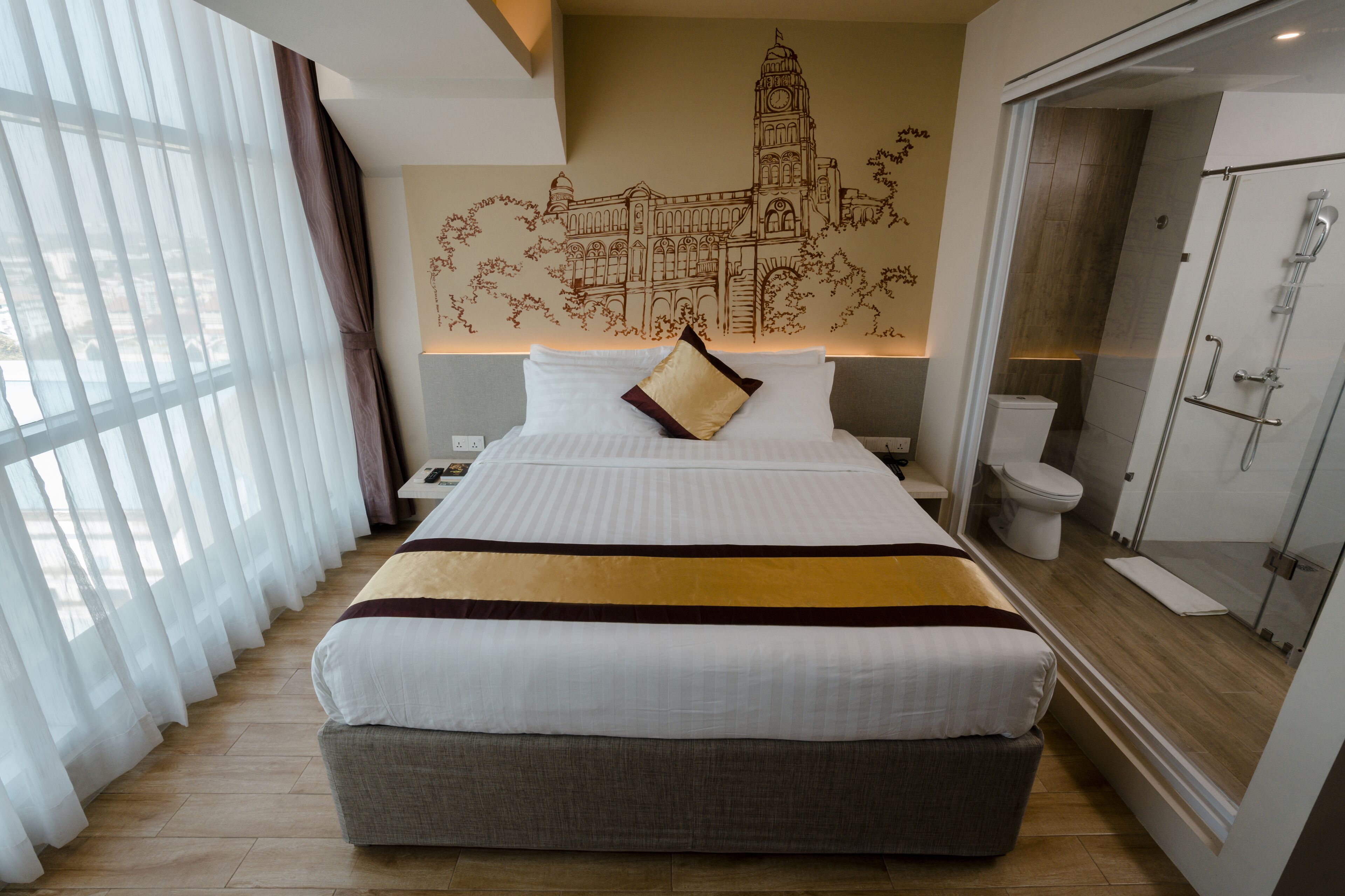 A hotel room with a large bed, a bathroom with a glass shower enclosure, and a mural of a building on the wall.