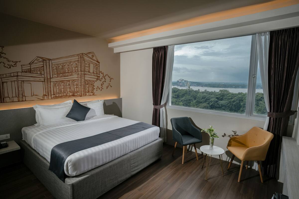 A modern hotel room with a large bed, a desk, two chairs, a view of a cityscape, and a wall mural.