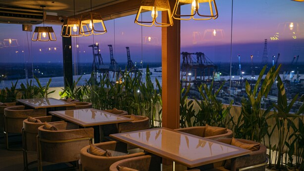 A restaurant with tables and chairs, large windows with a view of cranes and water, and hanging pendant lights.