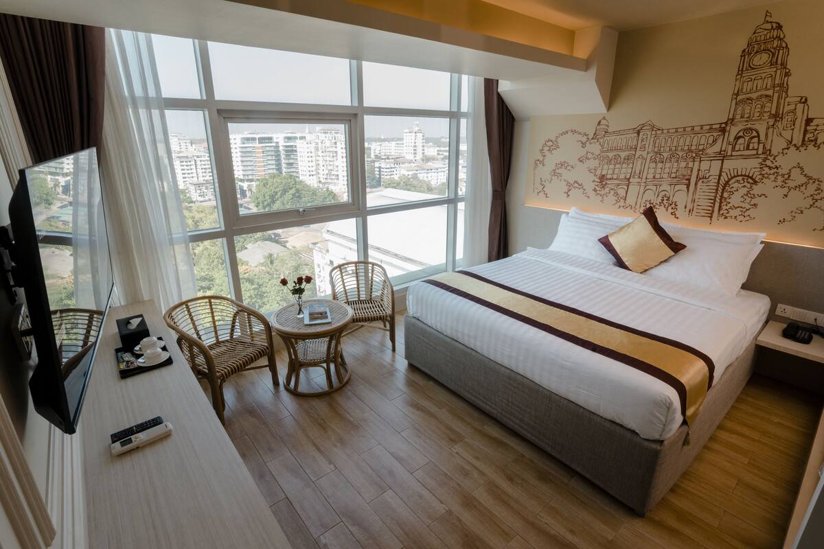 A hotel room with a large bed, a TV, a small table with a chair, and a view of the city through large windows.