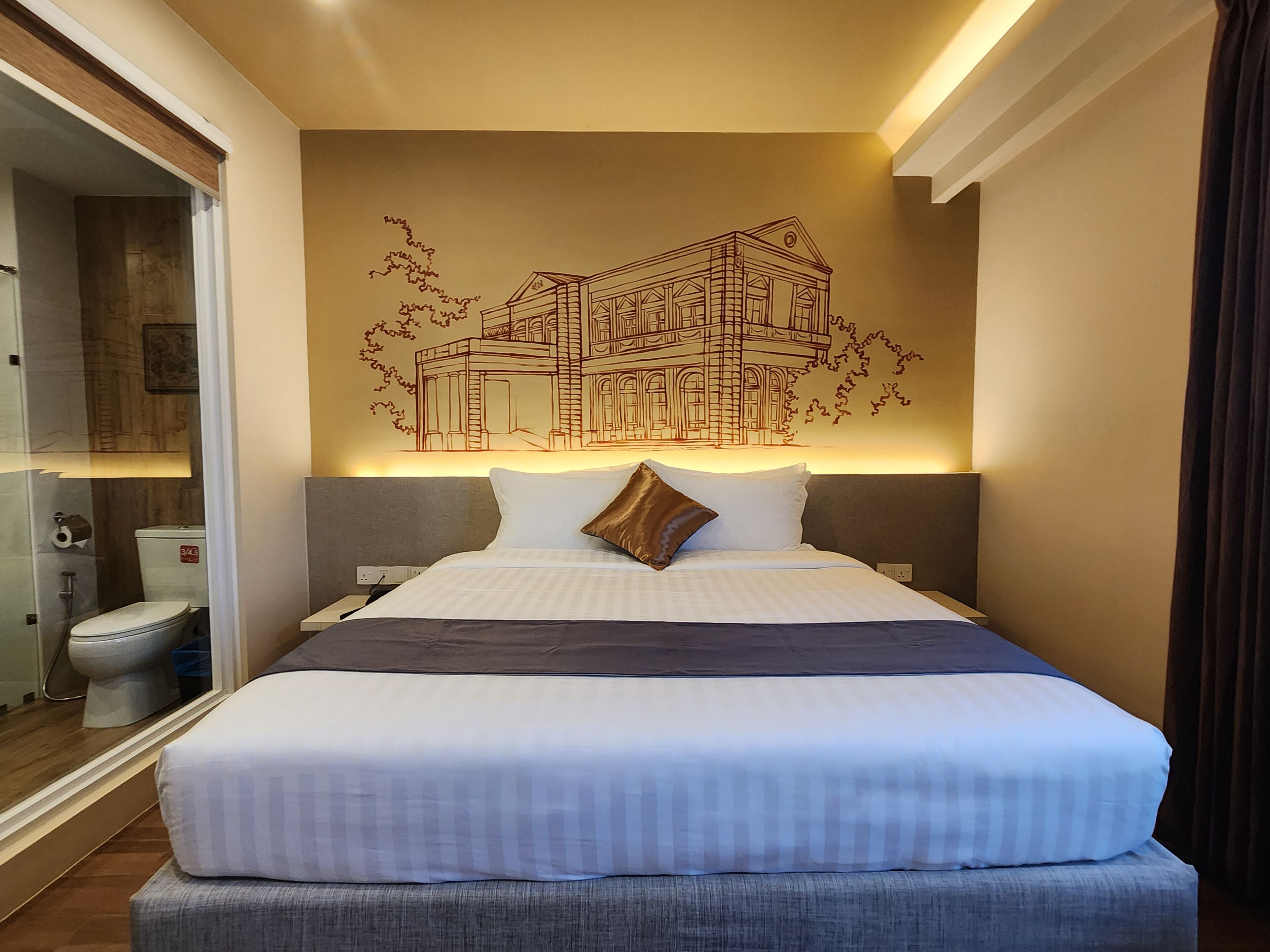 A hotel room with a large bed, a headboard, a wall-mounted artwork of a building, and a bathroom visible through an open door.