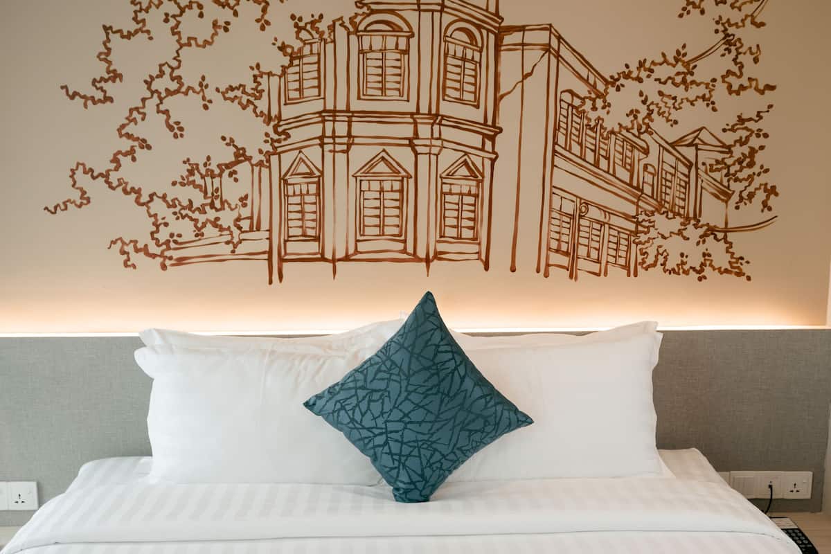 A hotel room with a bed, white bedding, a teal pillow, and a wall mural of a building with vines.