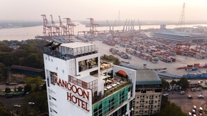 A high-rise hotel with multiple floors, a rooftop terrace, and a view of a port with shipping containers and cranes.