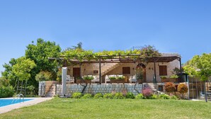 Un edificio de piedra con piscina, rodeado de vegetación y un cielo azul despejado.