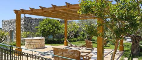 A wooden pergola with a stone bench and a table, surrounded by greenery and a stone wall.
