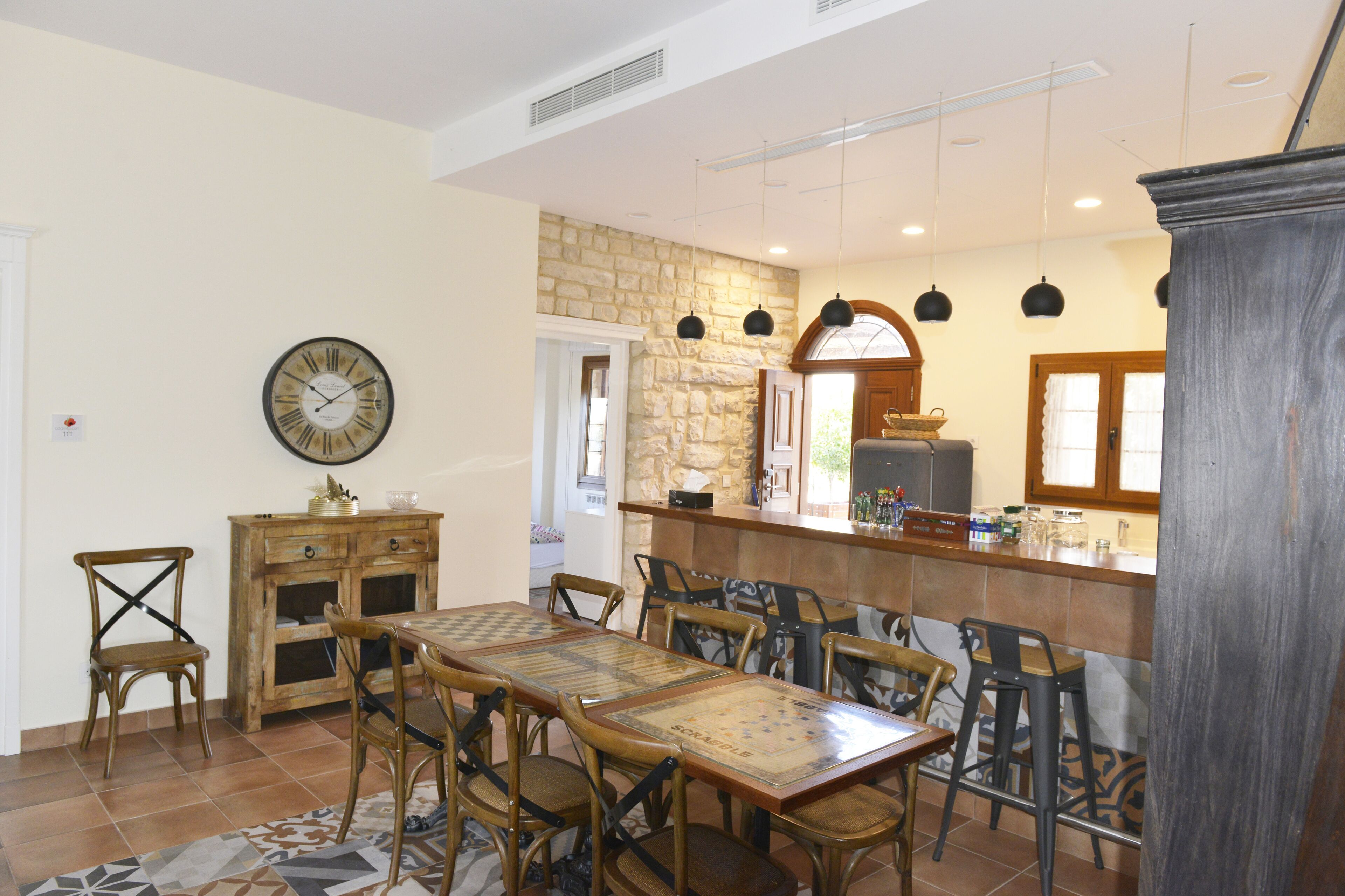 A dining area with a wooden table and chairs, a clock on the wall, and a kitchen area with a stone wall and hanging lights.