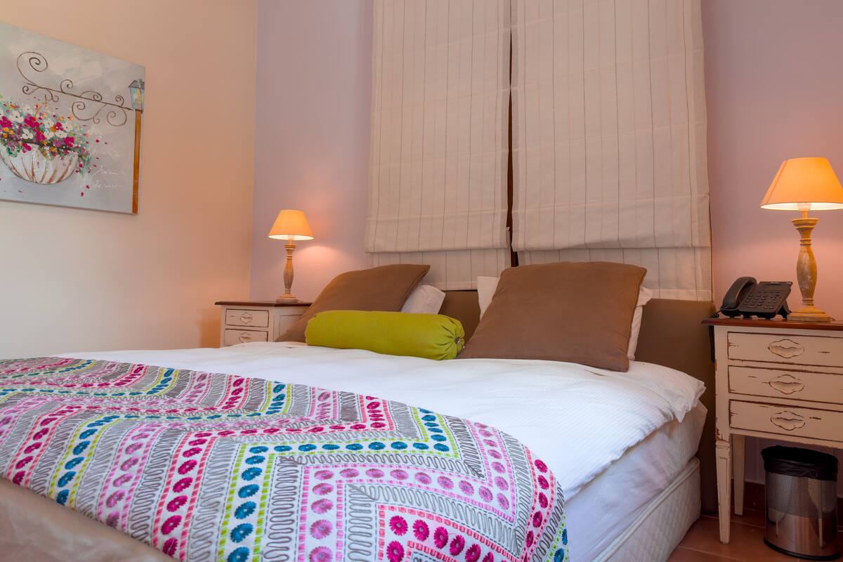A hotel room with a bed, two bedside tables, a lamp on each table, a trash can, and a painting on the wall.