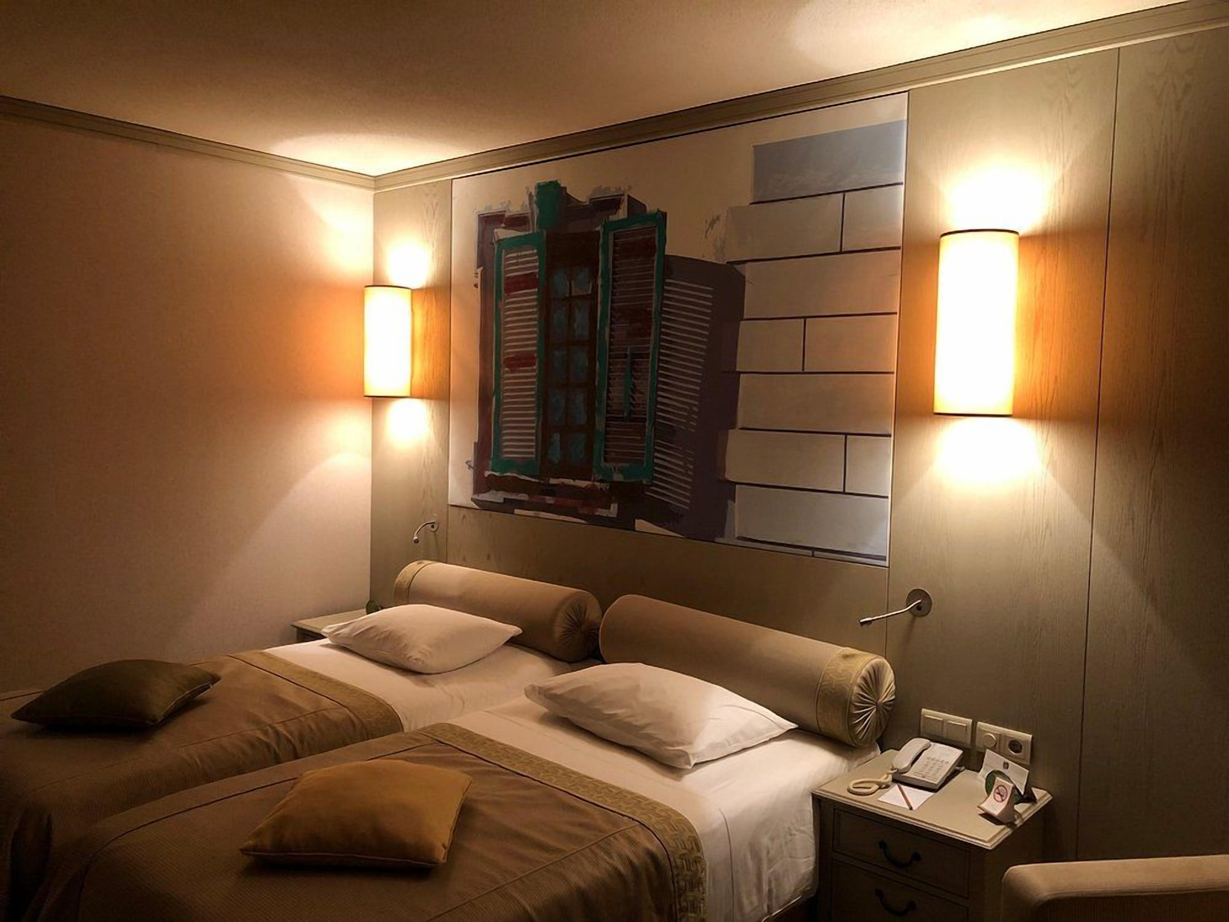 Habitación de hotel con cama, mesitas de noche, lámpara y una obra de arte en la pared.