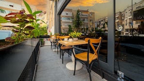 An outdoor café with tables and chairs, surrounded by potted plants and a cityscape view.