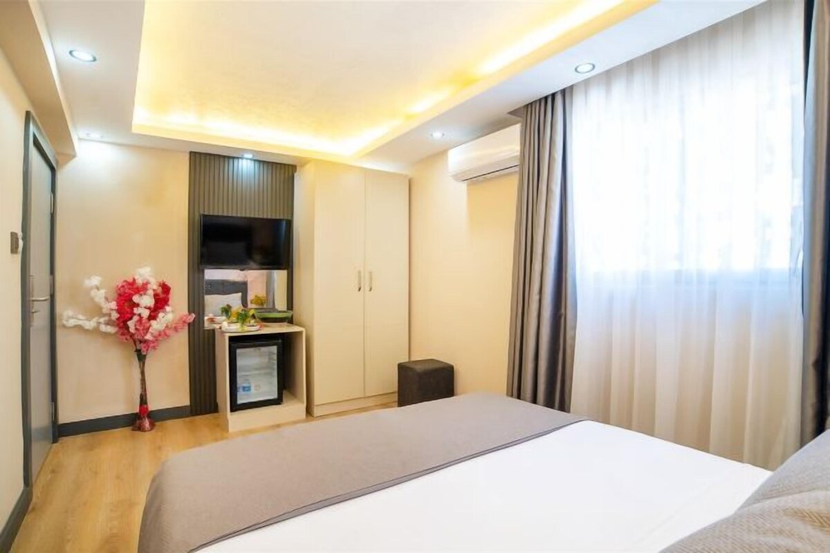 A modern hotel room with a large bed, a small desk, a TV, and a minibar.