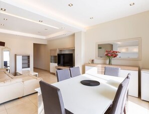 A modern living room with a sofa, dining table, and chairs. There is a TV, a mirror, and a kitchen area in the background.