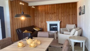 Una sala de estar acogedora con un muro de madera, una chimenea, un sofá de cuero negro, un sofá blanco y una mesa de comedor con tazas amarillas y una tetera.