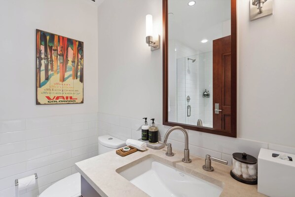 A modern bathroom with a large sink, a mirror, and a toilet. There is a painting on the wall and a shower visible in the background.