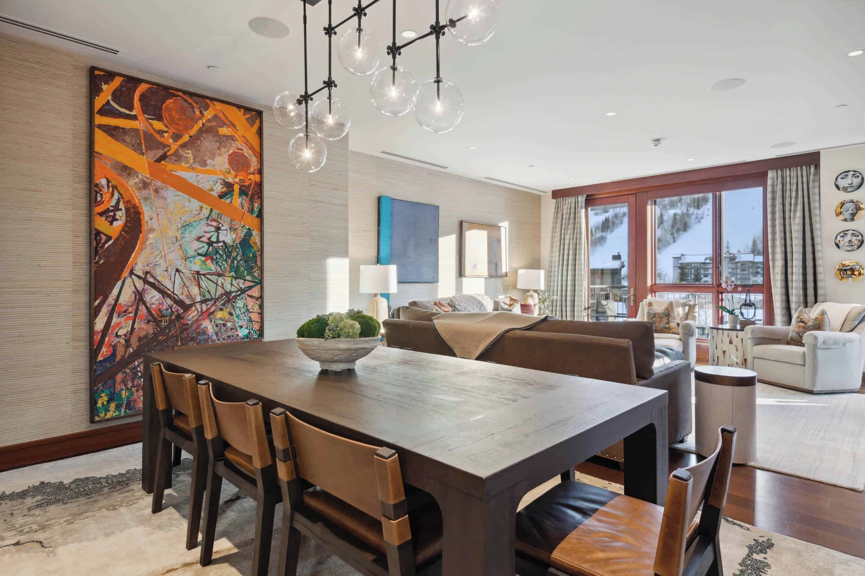 A modern dining area with a wooden table, chairs, and a large abstract painting. There is a sofa and a window with curtains in the background.