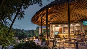 An outdoor seating area with a thatched roof structure, surrounded by greenery and overlooking a hilly landscape. - Phapok Eco Resort (Suan Phueng)